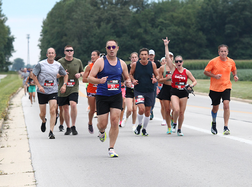 Runners in the Scouting America Half Marathon head down a road past cornfields in Bowling Green, Ohio, one cheering for the camera.